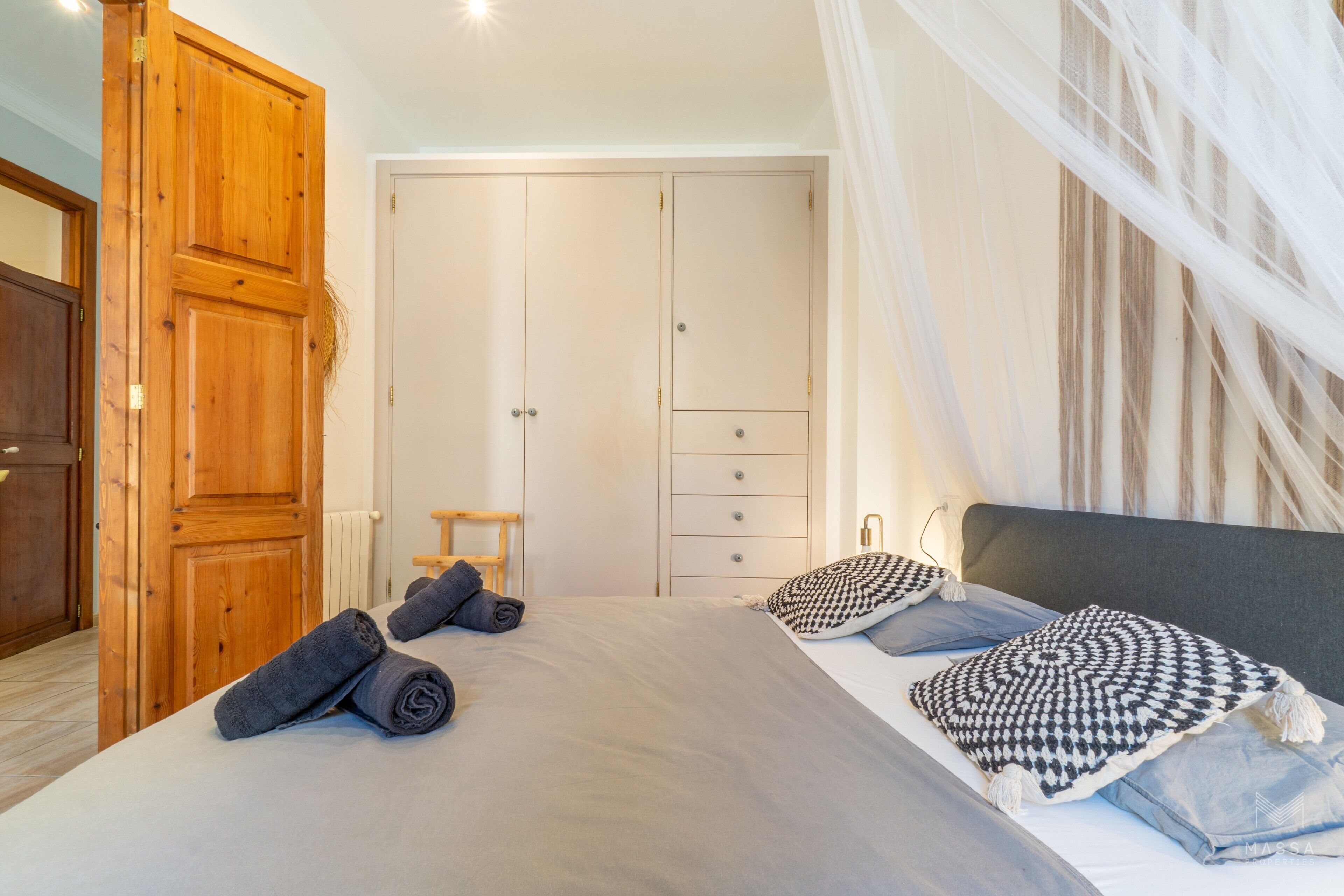A bedroom with a bed, a wooden wardrobe, a small stool, and a window with curtains.