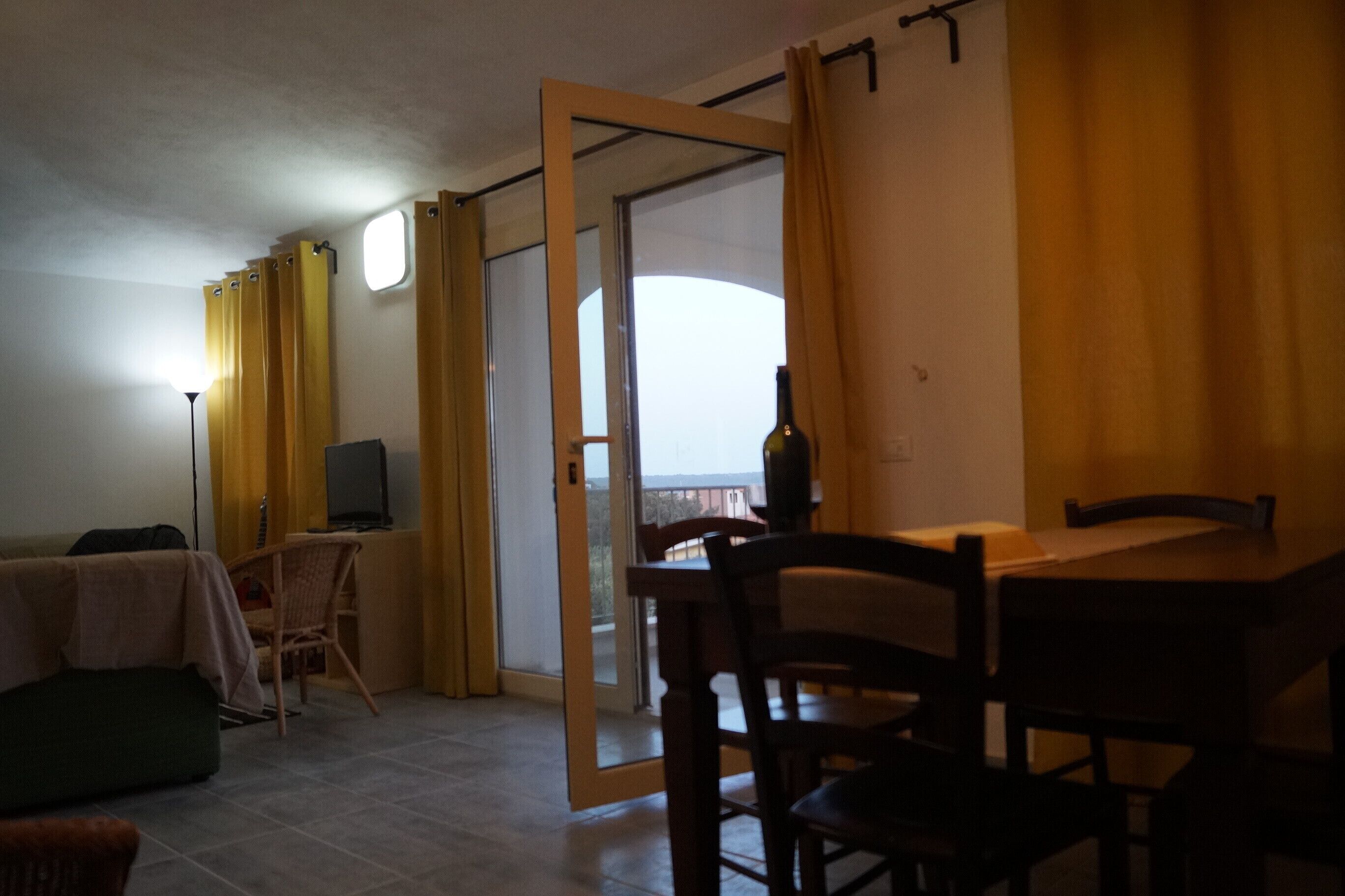 A room with a dining table and chairs, a sofa, a TV, and a bottle of wine on a table.
