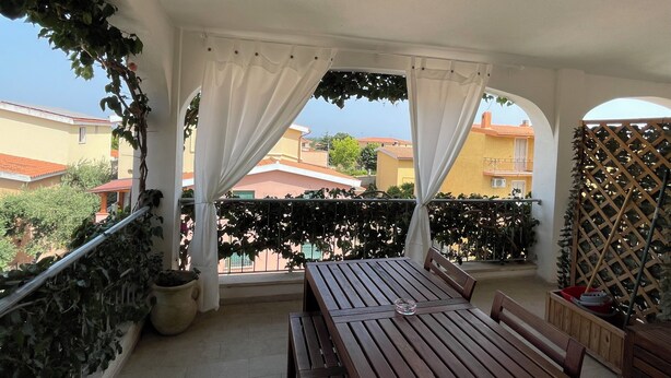 A balcony with a wooden table and benches, white curtains, and a view of neighboring buildings. - Sa Domo de Mannoi, home just minutes from Sardinia's most beautiful beaches (Orosei)