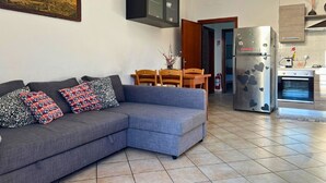 Ein modernes Wohnzimmer mit einem grauen Ecksofa, einem Holzesstisch und einer Küchenzeile inklusive Kühlschrank und Backofen.