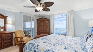 Un dormitorio con una cama grande, un aparador de madera, una silla, un ventilador de techo y vista al océano.