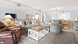Amplia sala de estar con sofá de cuero, una sección modular gris y una mesa de centro de madera. Hay un comedor con una mesa y sillas blancas.