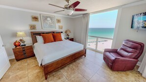 A hotel room with a bed, a ceiling fan, a view of the beach, a red leather chair, and artwork on the walls. - Beachfront Views from ST 1104 with Free Beach Service for 2! (Destin)