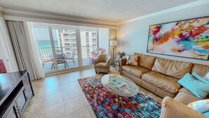 A living room with a sofa, a coffee table, a TV, and a balcony with a view of buildings and the sea. - Beachfront Views from ST 1104 with Free Beach Service for 2! (Destin)
