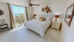 Een slaapkamer met een bed, een plafondventilator, een nachtkastje met een lamp, een decoratief wandobject en uitzicht op de oceaan.