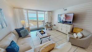 Een woonkamer met een bank, fauteuil, televisie en een balkon met uitzicht op zee.