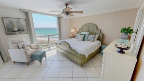 Una camera da letto con un letto, una sedia, un comodino, una lampada, una ventola a soffitto, vista sulla spiaggia e un quadro appeso al muro.