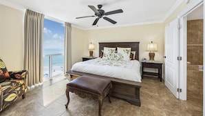 Ein Hotelzimmer mit einem Bett, zwei Nachttischen, einer Bank, einem Deckenventilator und Blick aufs Meer, sowie einer Tür zu einem Badezimmer.