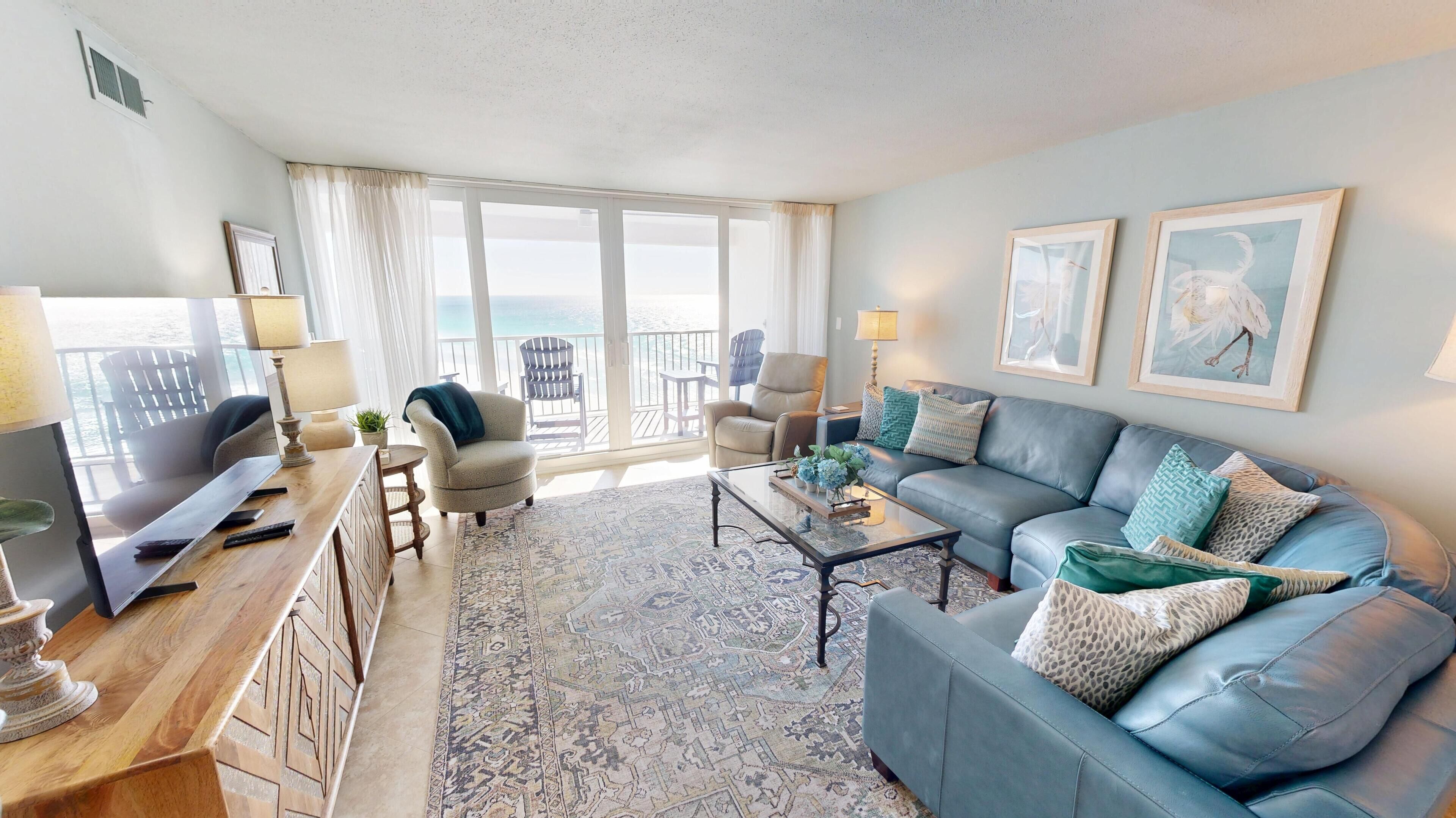 A spacious living room with a large sofa, a coffee table, and a TV on a wooden desk. There are two chairs and a small table with a lamp. The room has a large window with a view of the ocean.