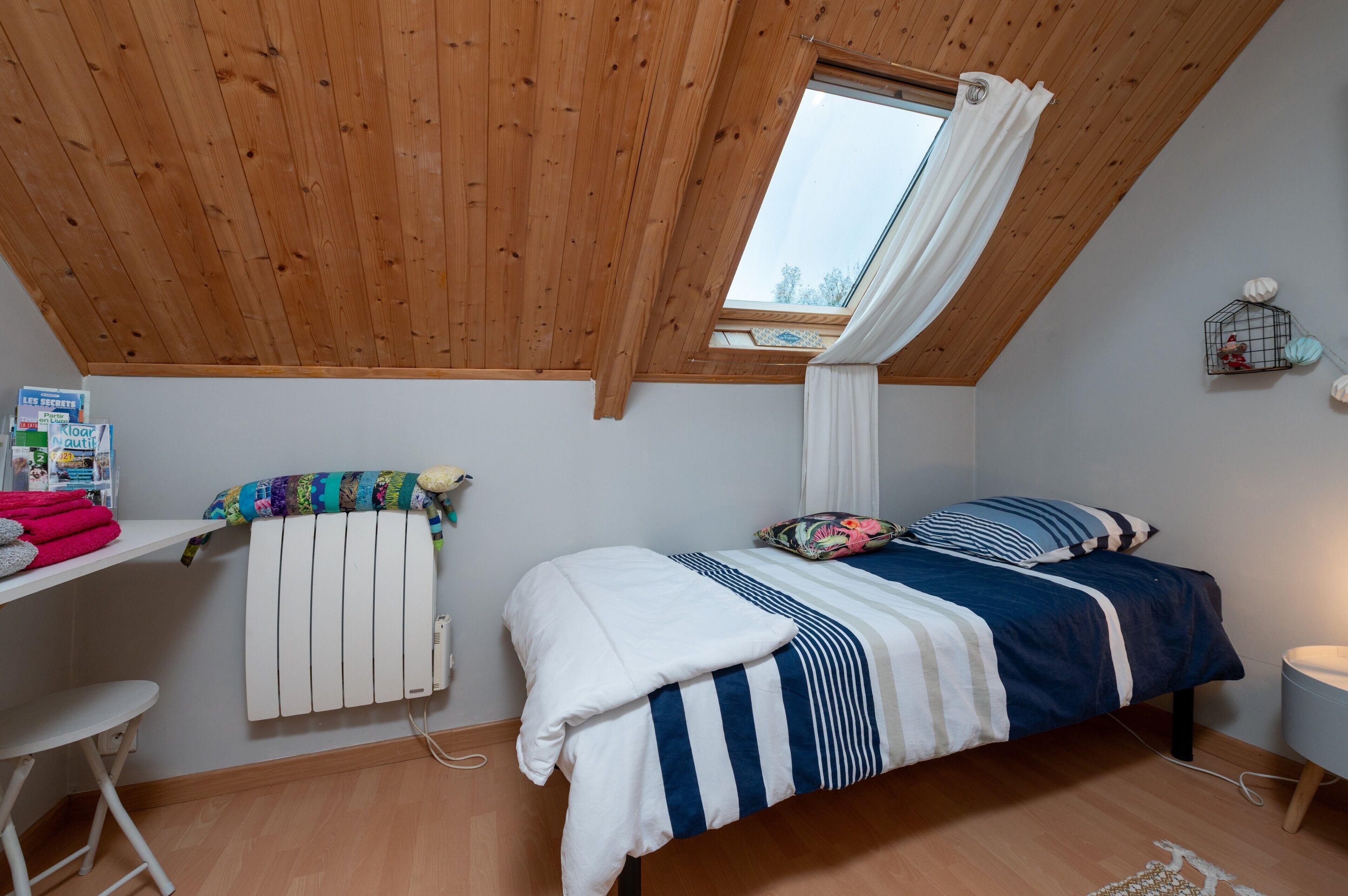 Une chambre avec un plafond en pente, une fenêtre avec des rideaux, un lit avec une literie à rayures, un radiateur et une étagère avec divers objets.