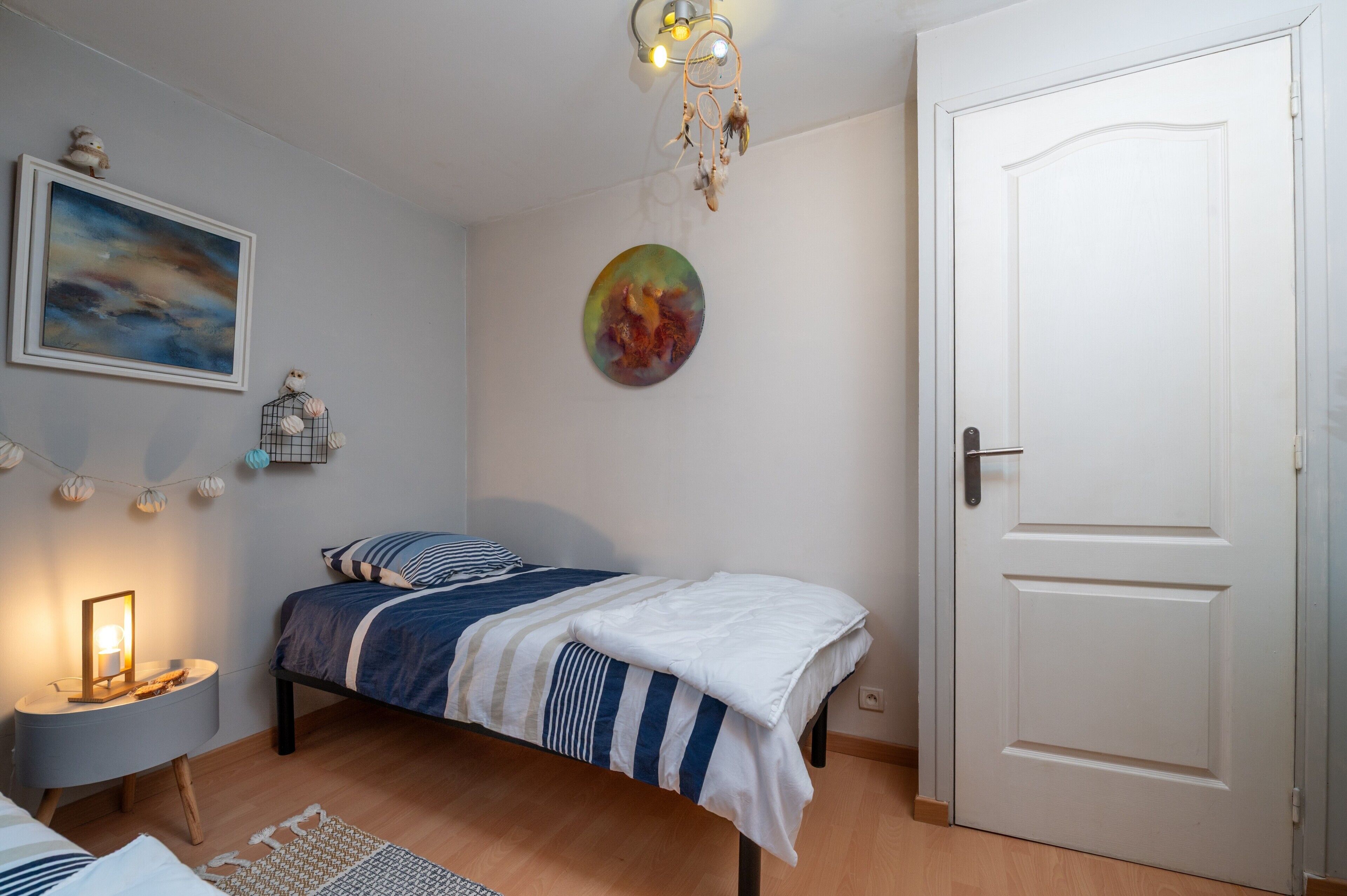A bedroom with a bed, a nightstand, a wall-mounted picture, a door, and a ceiling light fixture.