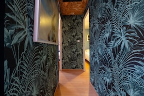 A hallway with a tropical leaf pattern wallpaper, a framed picture on the left, and a wooden floor.