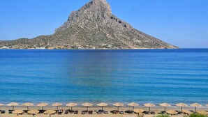 A beach with umbrellas and lounge chairs, a rocky island in the background, and clear blue water. - Seafront Casa Azul Villa - Getaway in Kalymnos (Mirties)