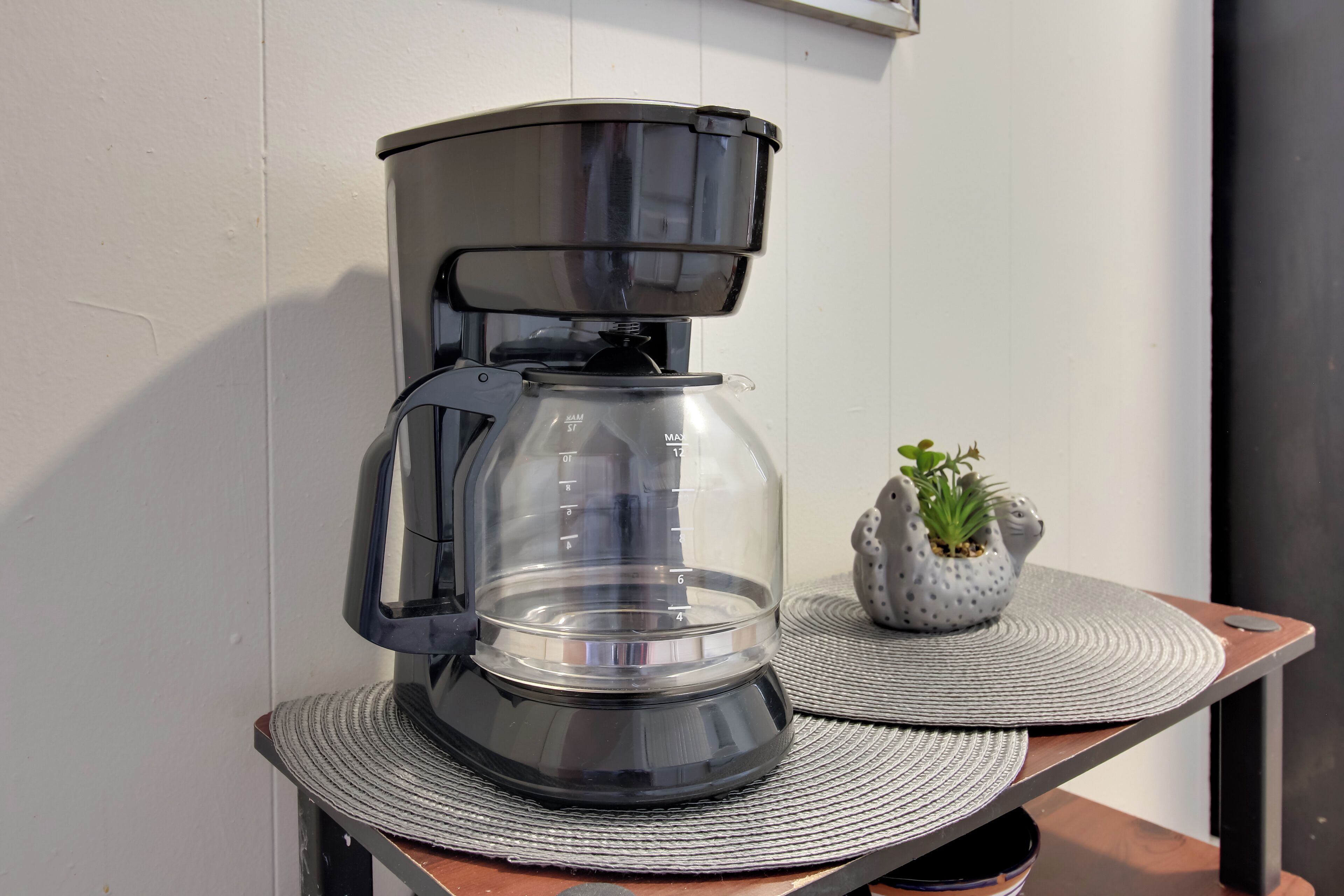 Une machine à café est posée sur une table, accompagnée d’un set de table, d’une petite plante en pot et d’un luminaire fixé au mur.