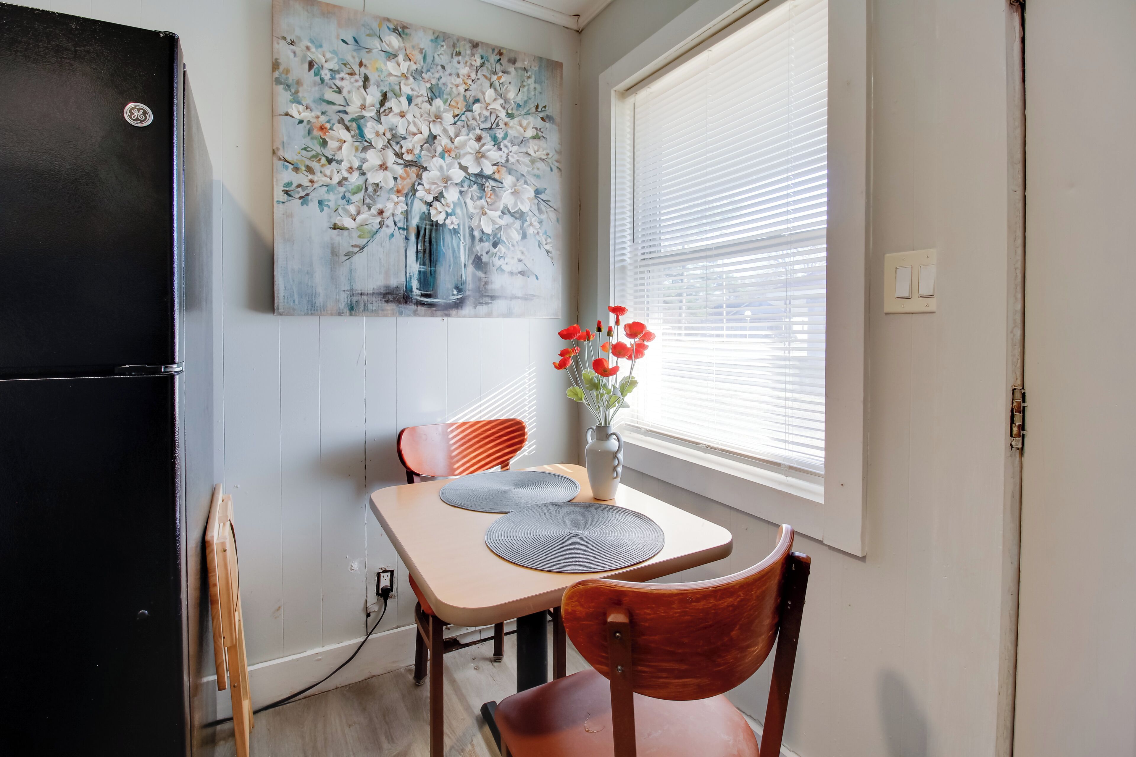 A dining area with a table, chairs, a vase with flowers, a painting on the wall, and a window with blinds.