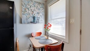 A dining area with a table, chairs, a vase with flowers, a painting on the wall, and a window with blinds. - Cozy Huntsville Studio Near Lowe Mill (Huntsville)