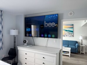 A hotel room with a large flat-screen TV displaying a live TV channel, a white dresser, a black chair, a small table with a lamp, and a painting of a beach scene. - Amazing Ocean Front 2BR 2 BA - Camelot 911 (Myrtle Beach)