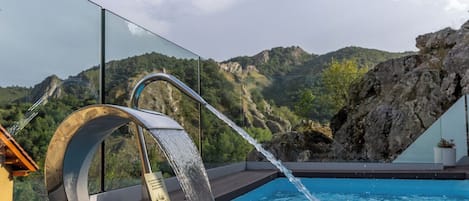 A modern outdoor pool with a waterfall feature, surrounded by a glass railing and overlooking a mountainous landscape.