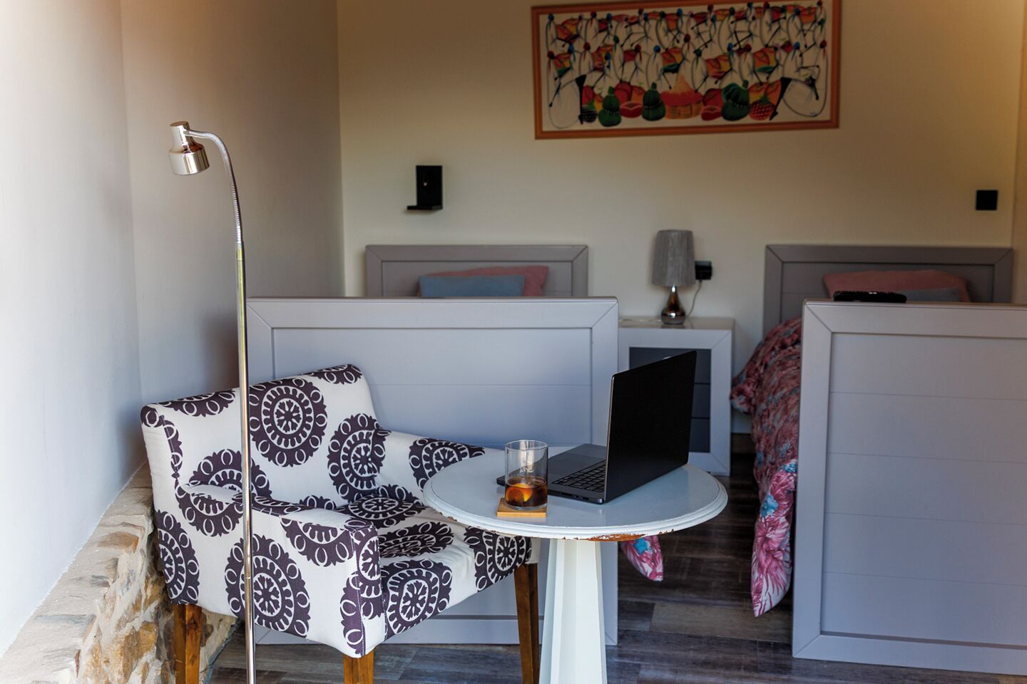 Quarto de hotel moderno com uma poltrona estofada, uma mesinha redonda com um laptop e um copo de bebida, e um quadro abstrato colorido na parede.