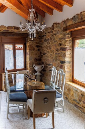 A dining area with a glass table surrounded by white chairs, a chandelier, stone walls, and a window with curtains. - El Refugio II de Verdiago (Cremenes)