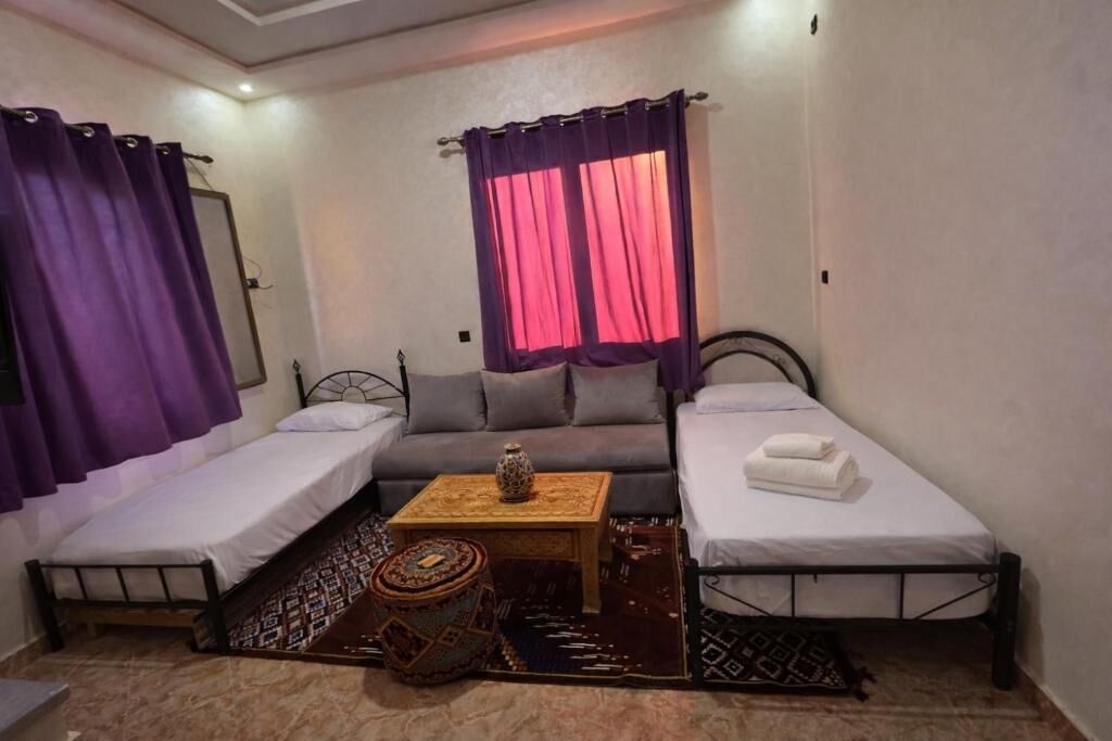 A small, well-lit room with two single beds, a sofa, a small table, and a patterned rug.
