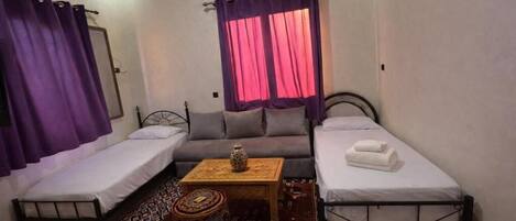 A small, well-lit room with two single beds, a sofa, a small table, and a patterned rug.