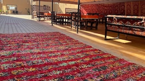 Un espace extérieur abrité, aménagé pour les repas, avec des tables et des chaises, une moquette à motifs et un bâtiment en arrière-plan.