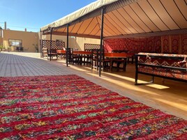 Un área de comedor al aire libre techada, con mesas y sillas, una alfombra estampada y un edificio al fondo.