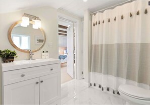 A bathroom with a white vanity, a round mirror with lights, a toilet, and a shower curtain with tassels. - Anchor’d in Hampton : Coastal Getaway Steps to Beach (Hampton)