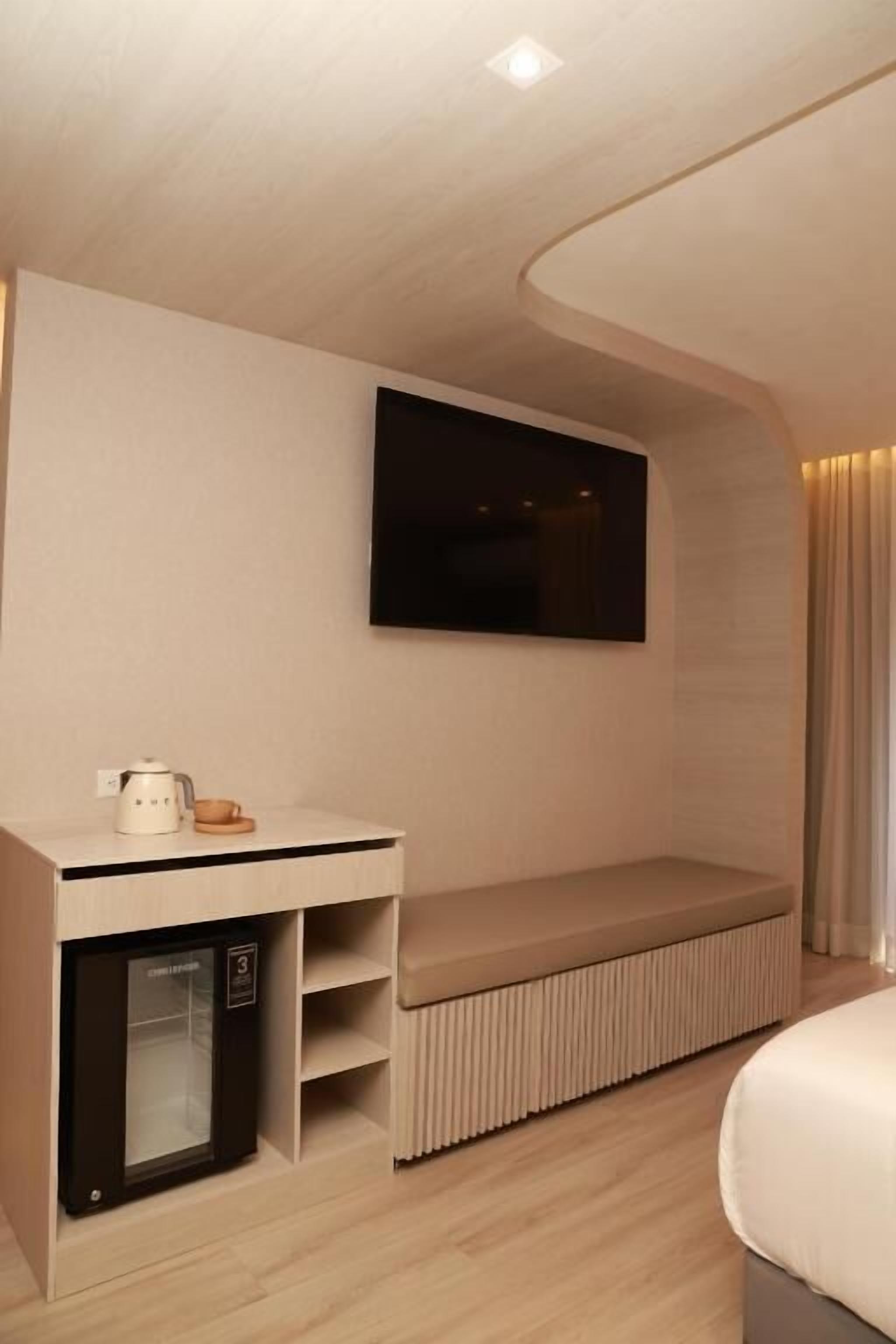 A modern hotel room with a built-in TV, a mini-fridge, a seating area, and a bed.