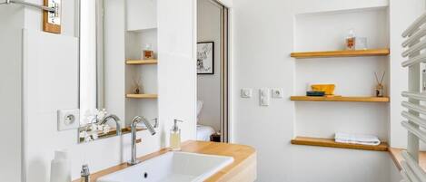 A modern bathroom with a wooden vanity, a white sink, and a mirror. There are wooden shelves above the sink and a door leading to another room.