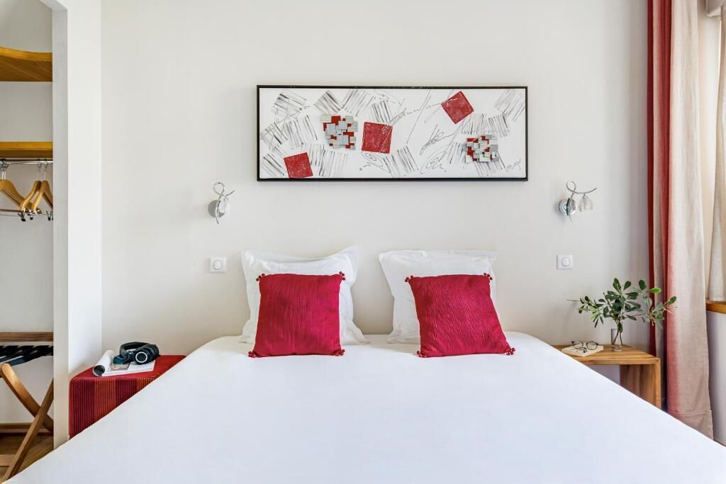 Una cama bien hecha con dos almohadas rojas, una obra de arte abstracta enmarcada encima y una mesita de noche con una planta y un par de gafas.