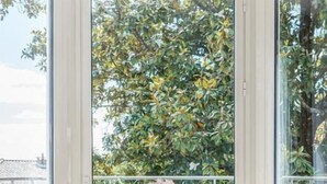 Ein Eckzimmer mit einem Holztisch und zwei gemusterten Stühlen, ein großes Fenster mit Blick auf Grünflächen und ein weißes Geländer draußen.