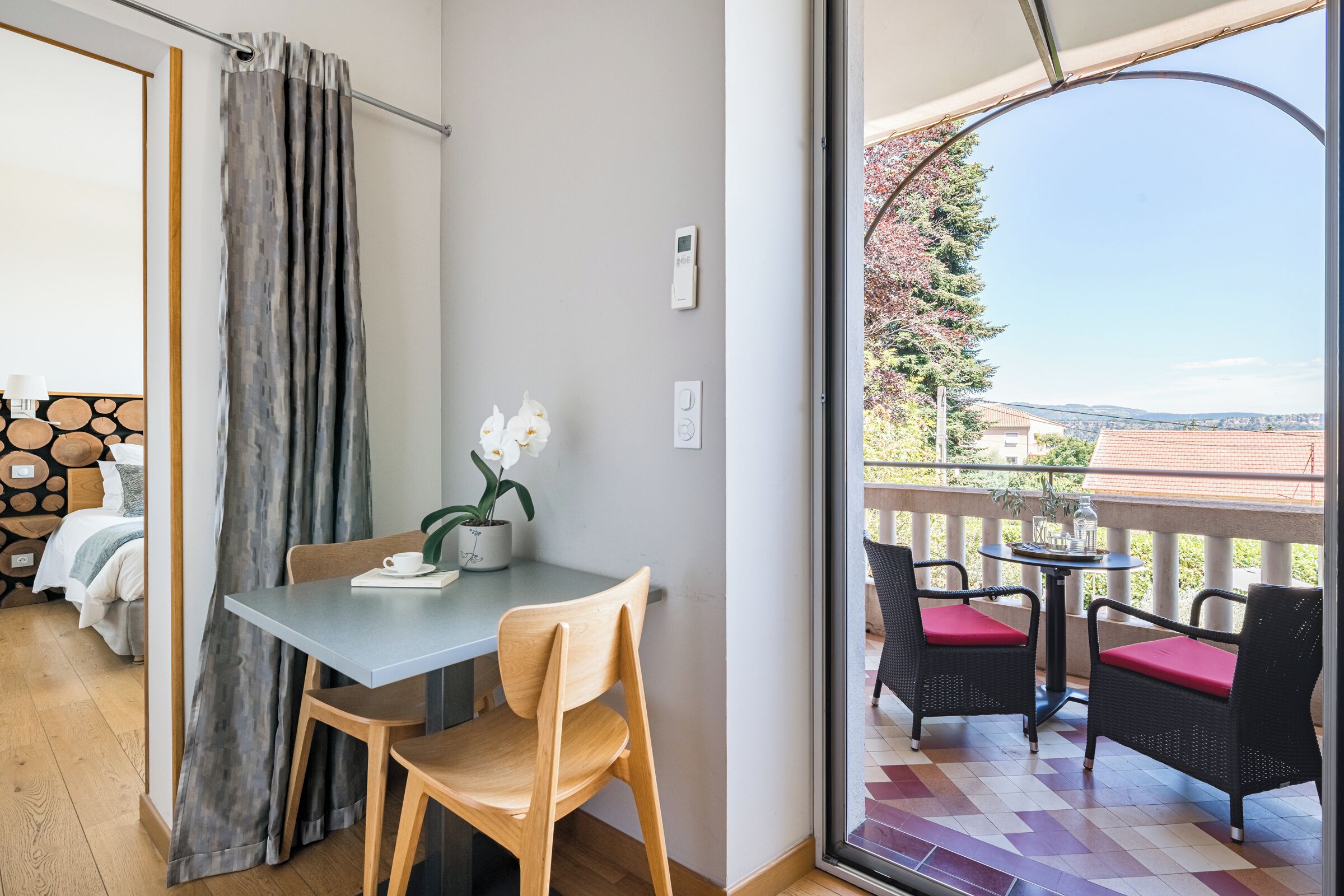 A room with a balcony, a dining table with chairs, a bed with a headboard, and a window with curtains.