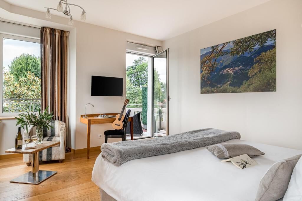 A modern bedroom with a large bed, a desk with a guitar, a TV, and a balcony with a view of trees.