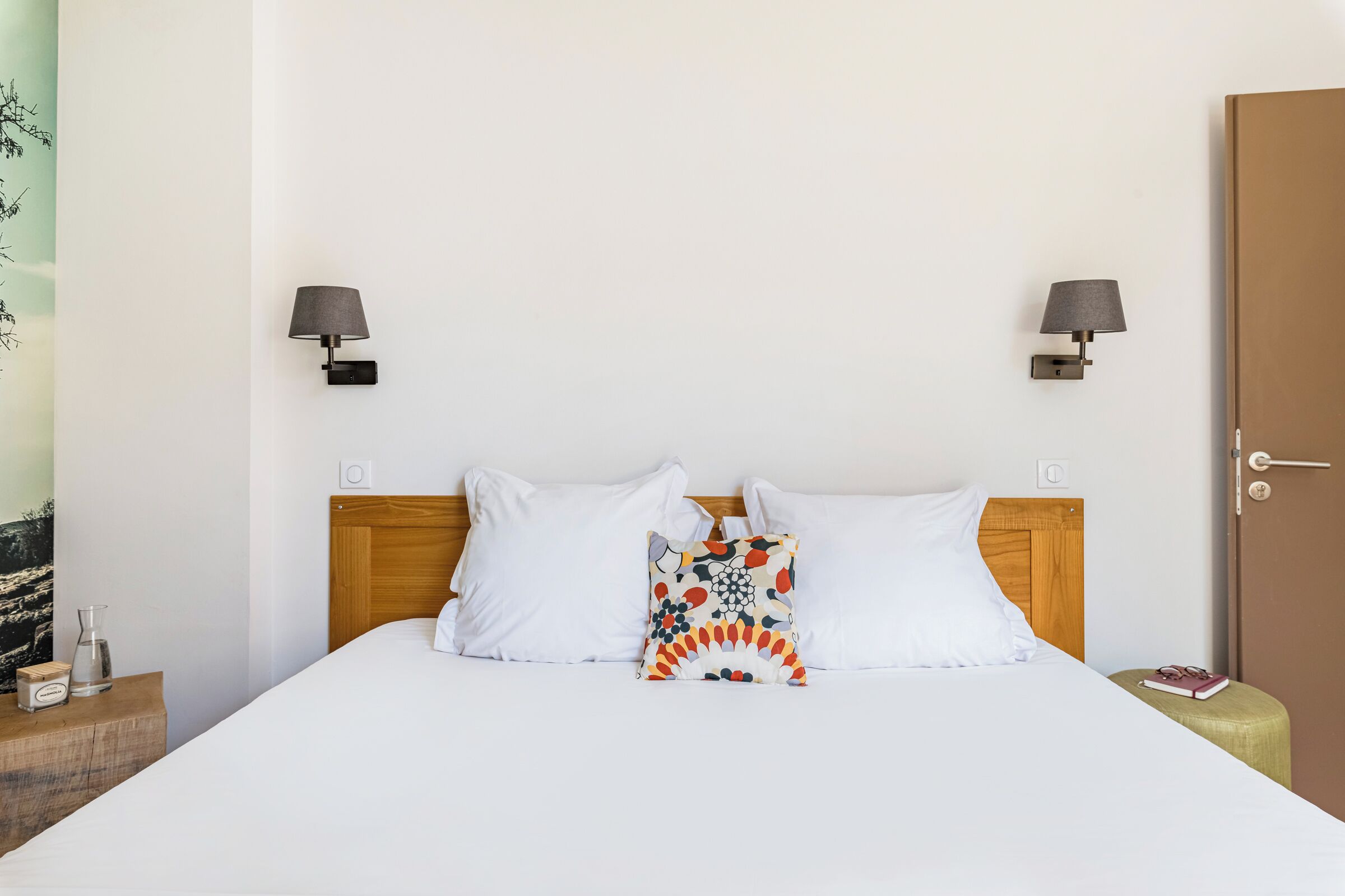 A hotel room with a wooden headboard, white bedding, a colorful pillow, two wall-mounted lamps, a door, and a small bedside table with a book and a glass.