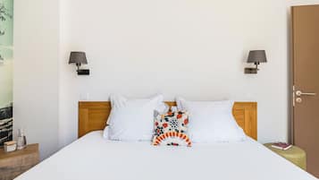 A hotel room with a wooden headboard, white bedding, a colorful pillow, two wall-mounted lamps, a door, and a small bedside table with a book and a glass.