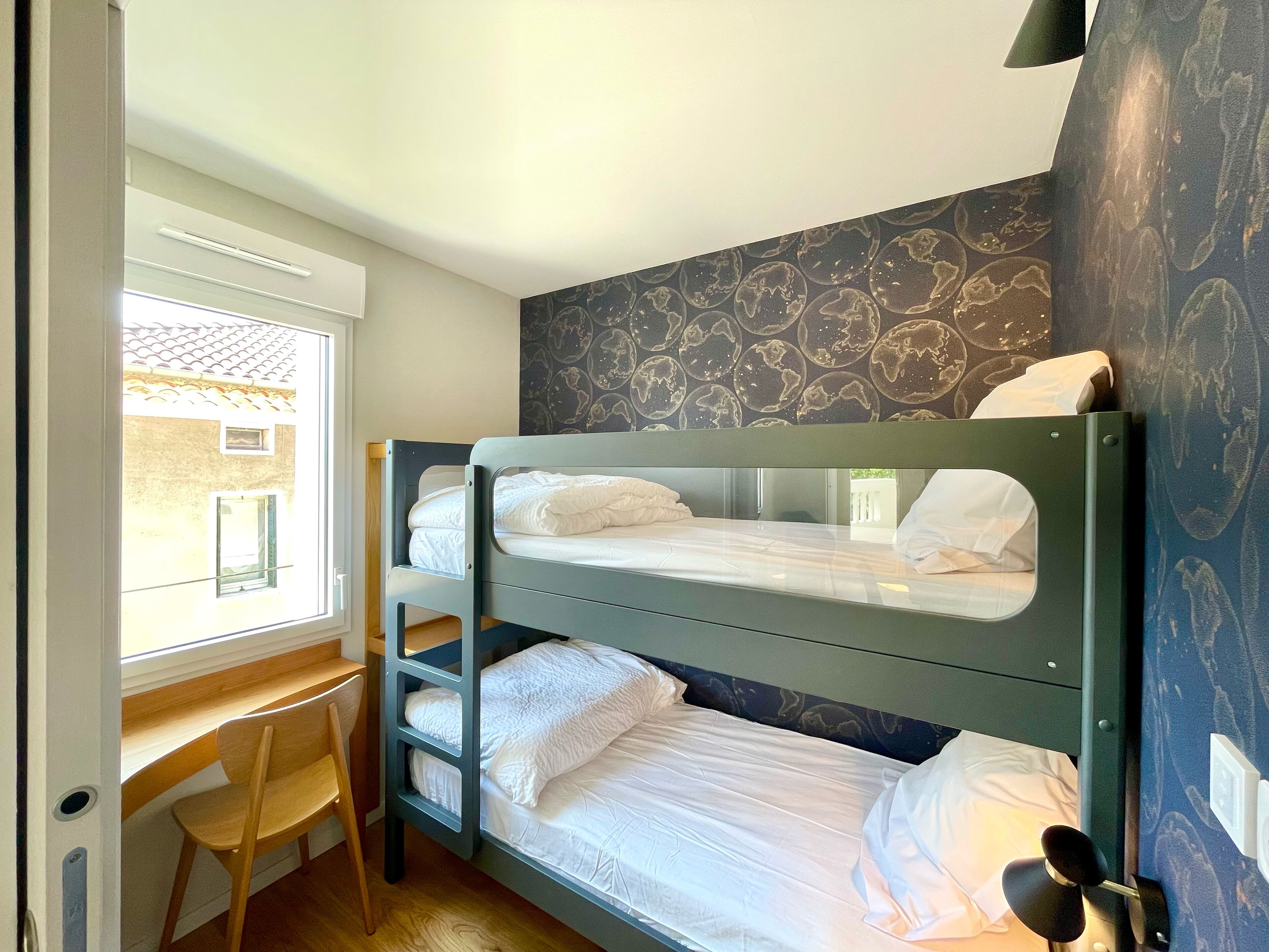 Quarto com beliche, escrivaninha, cadeira, janela e parede estampada.