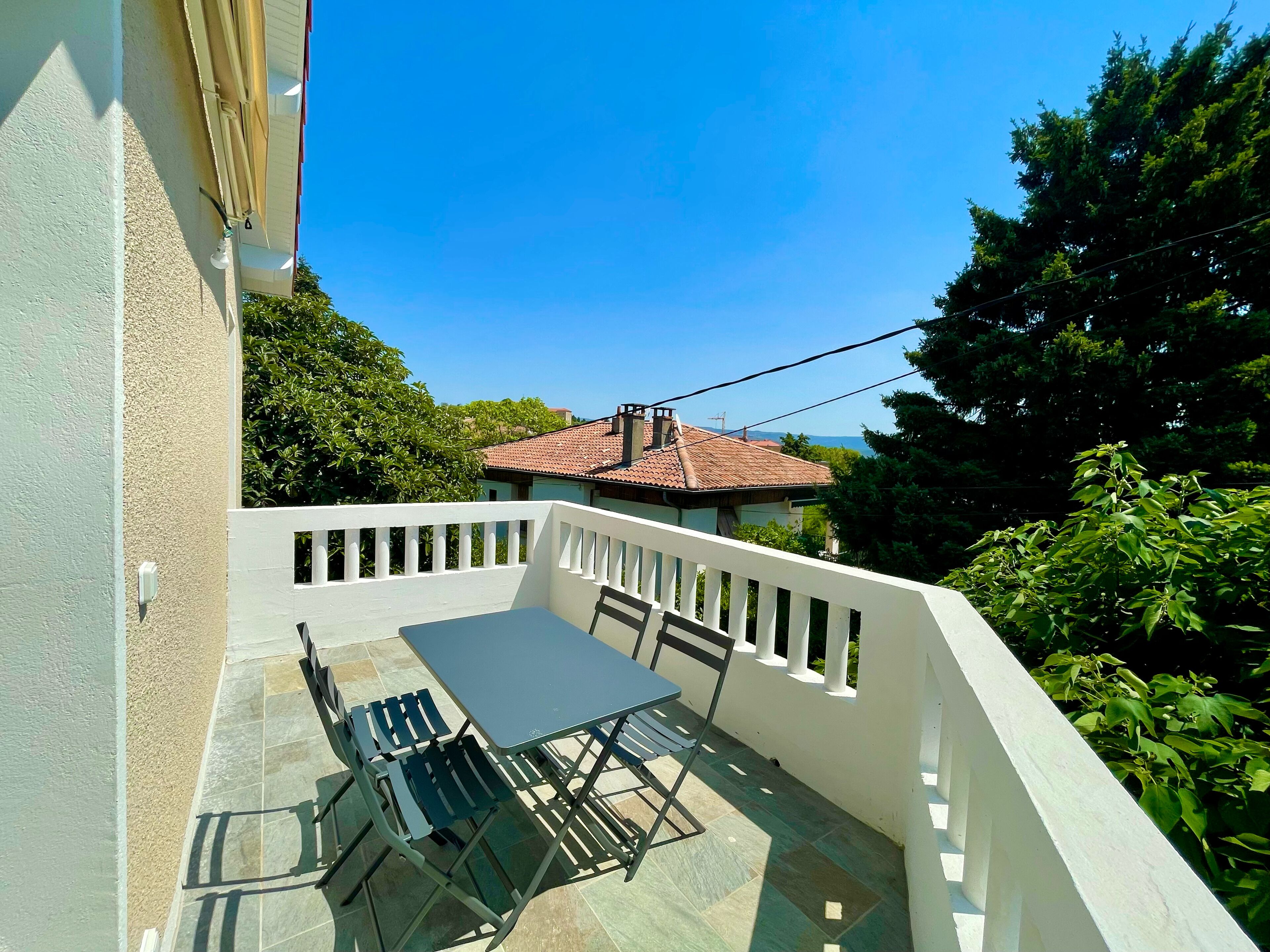 A balcony with a table and chairs, a white railing, and a view of a red-roofed building in the background.