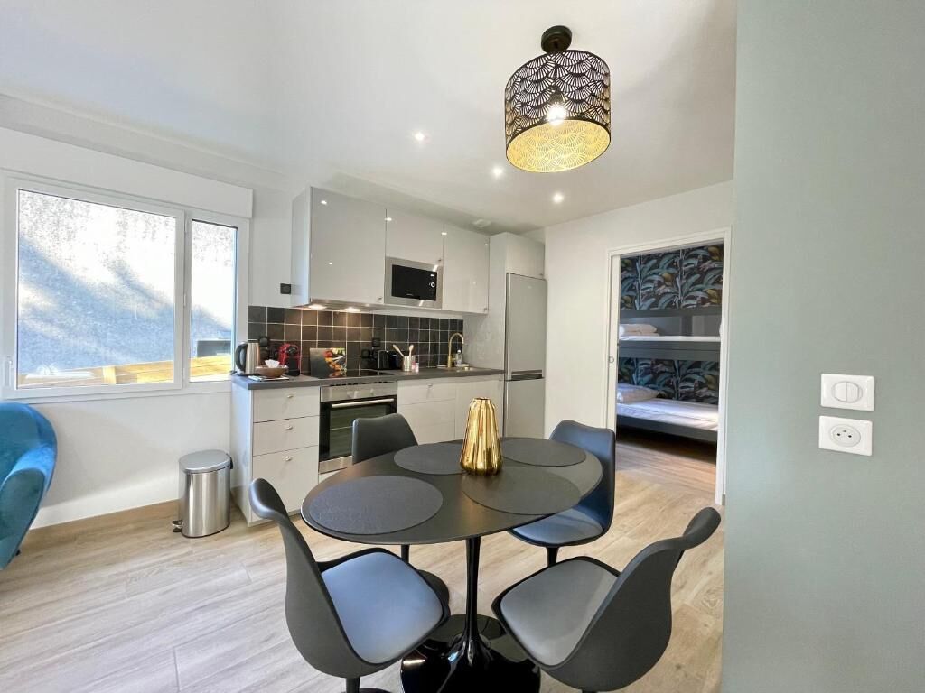 A modern kitchen with a dining area, featuring a round table with chairs, a hanging light fixture, and a view of the outdoors.