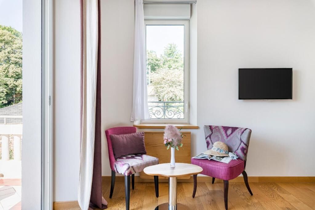 Een kamer met een groot raam, twee stoelen, een klein tafeltje en een flatscreen televisie aan de muur.