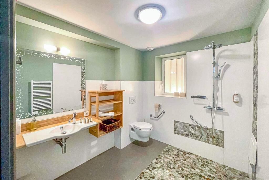 Un baño con lavamanos, inodoro y ducha. Hay un espejo sobre el lavamanos y una ventana sobre el inodoro.