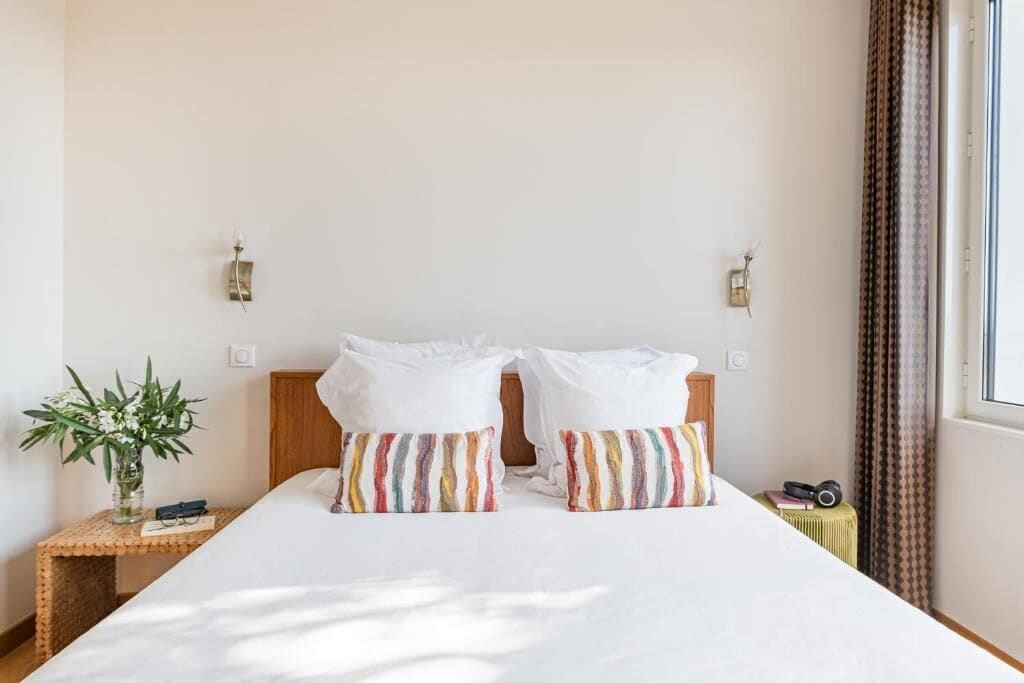 Hotelkamer met een groot bed, twee kussens, een nachtkastje met een plant, een camera en een raam met gordijnen.