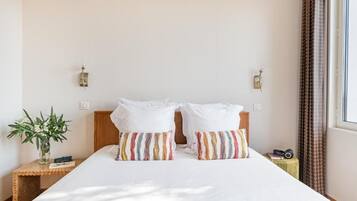 Habitación de hotel con una cama grande, dos almohadas, una mesita de noche con una planta, una cámara y un ventanal con cortinas.