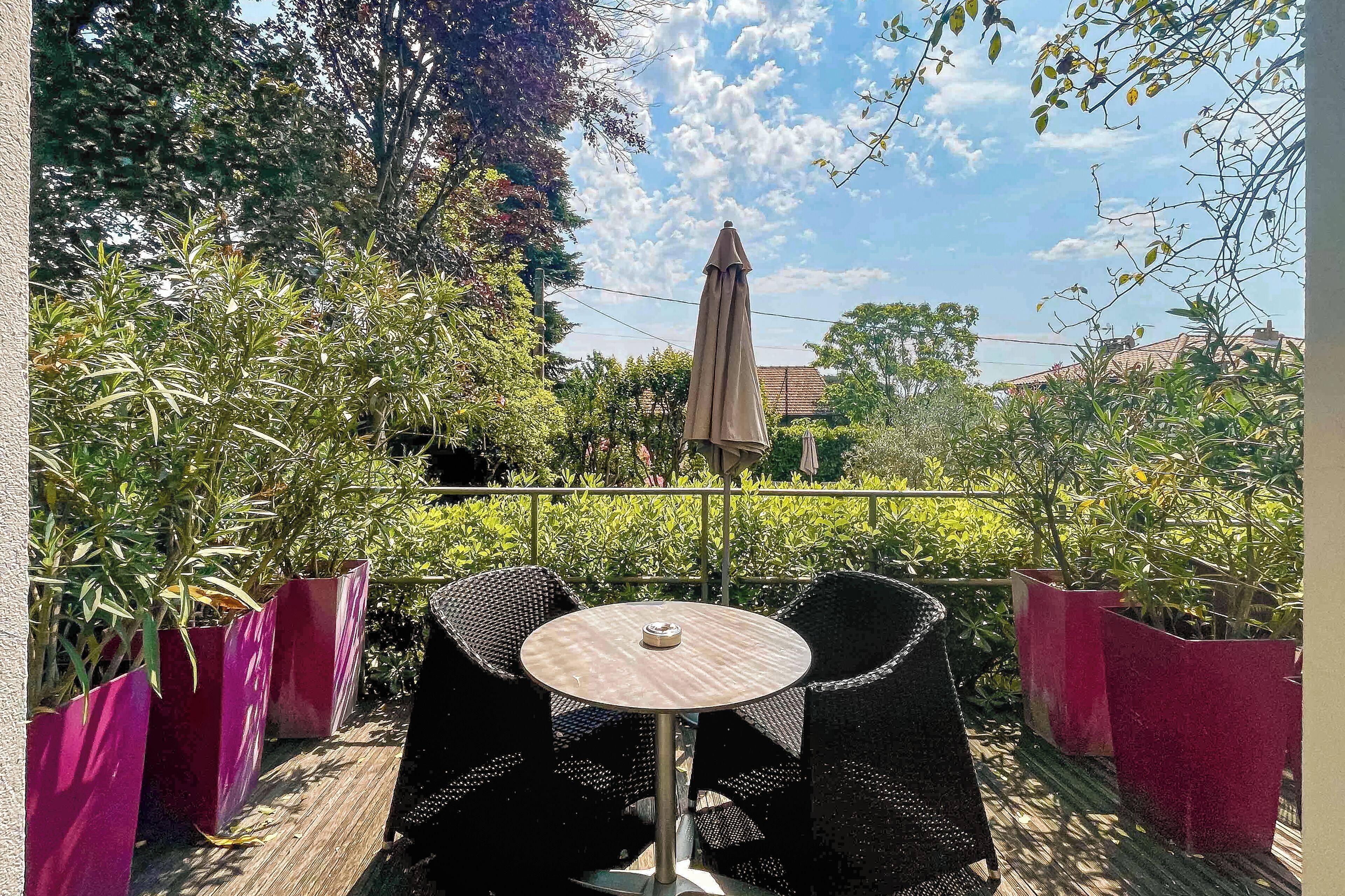 An outdoor patio with wicker chairs and a round table, surrounded by potted plants and a large umbrella.