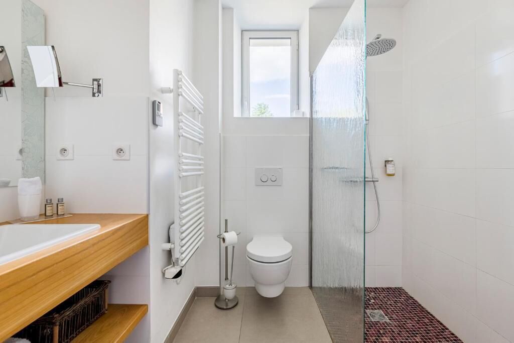 Een moderne badkamer met een houten wastafelmeubel, een toilet, een douchecabine met glazen wanden en een raam boven het toilet.