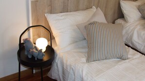 Una cama bien hecha con ropa de cama blanca y almohadas a rayas, una pequeña silla negra con una lámpara y peluches, y un cabecero de madera.