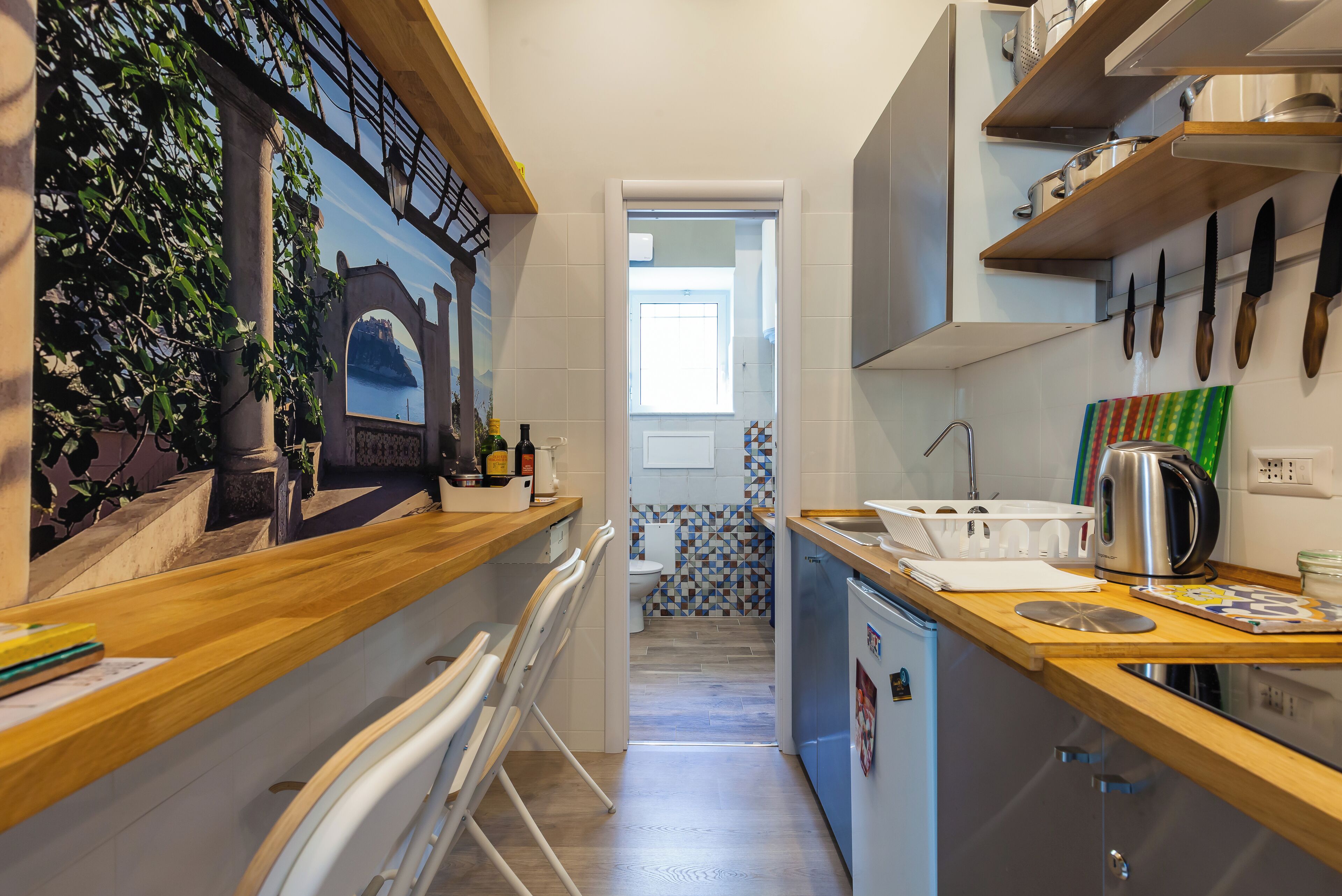 A narrow kitchen with a wooden countertop, a small sink, and a refrigerator. There is a window with a view of a building and a door leading to a bathroom with a tiled wall.