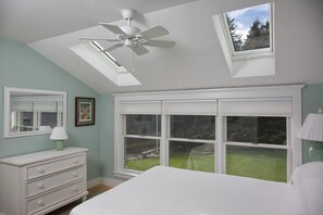 Une chambre à coucher comprenant un lit, une commode, un ventilateur de plafond, un puits de lumière et une grande fenêtre donnant sur une cour verdoyante.