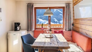 A wooden dining table with a wine bottle and glasses, a chair, a small refrigerator, and a view of snow-covered mountains through a window.